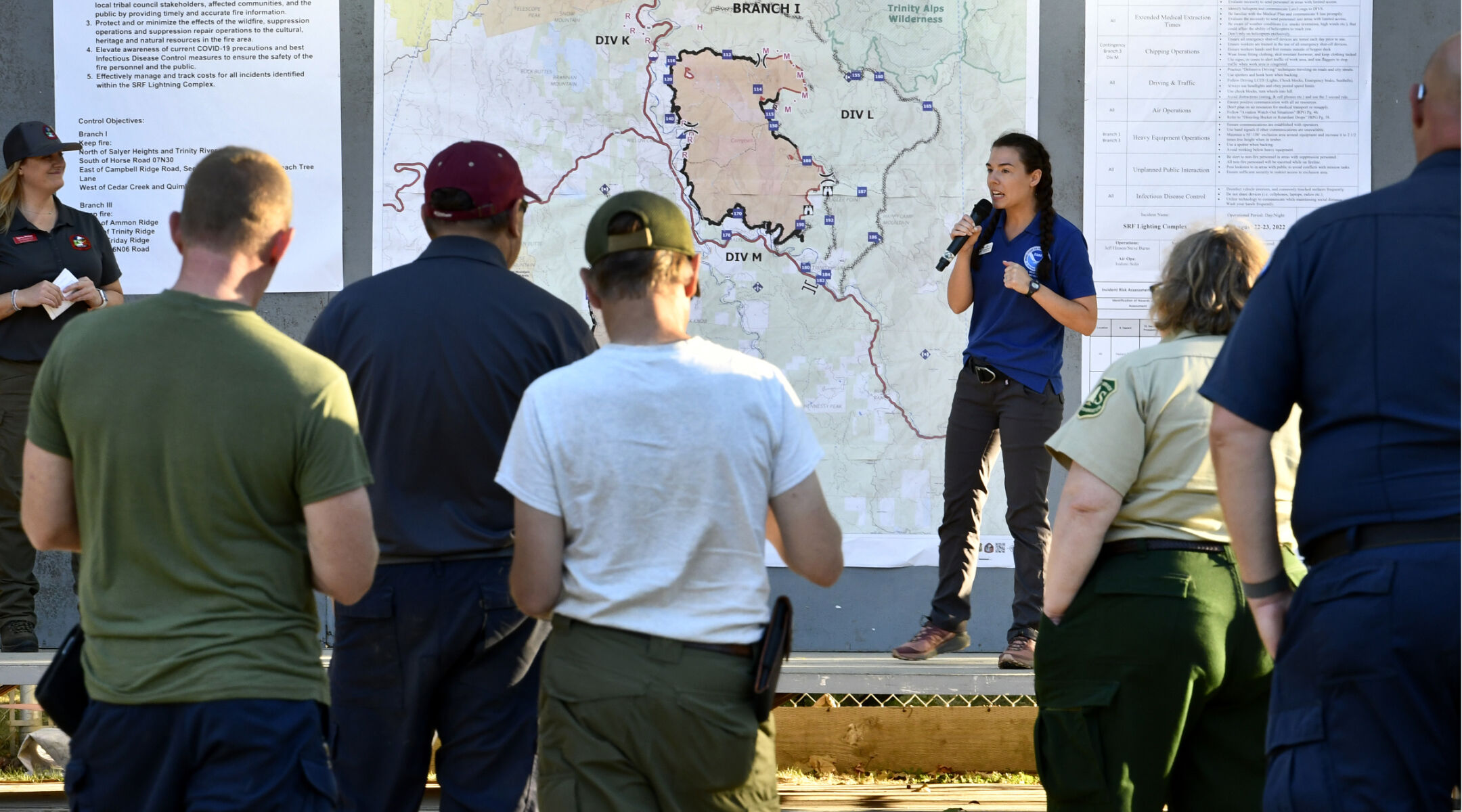 A National Weather Service meteorologist is briefing a crowd of people outdoors in front of a large map and charts of data.